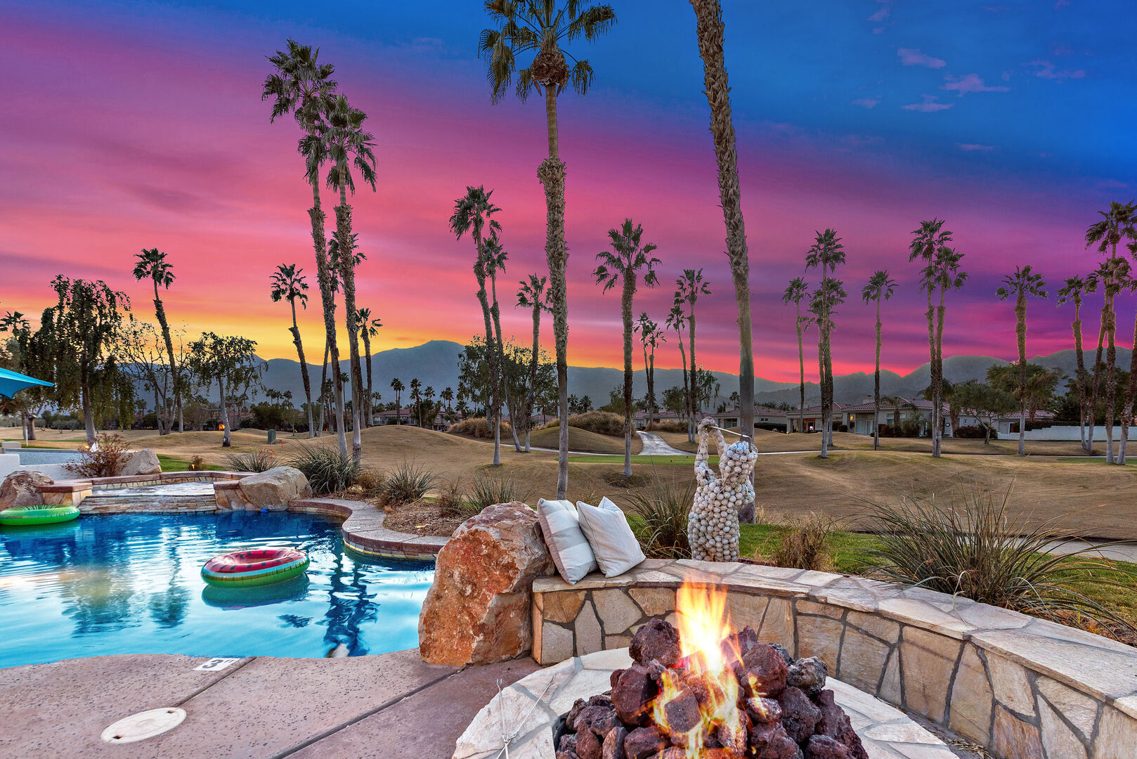 A desert pool and stone fire pit beneath palm trees at sunset
